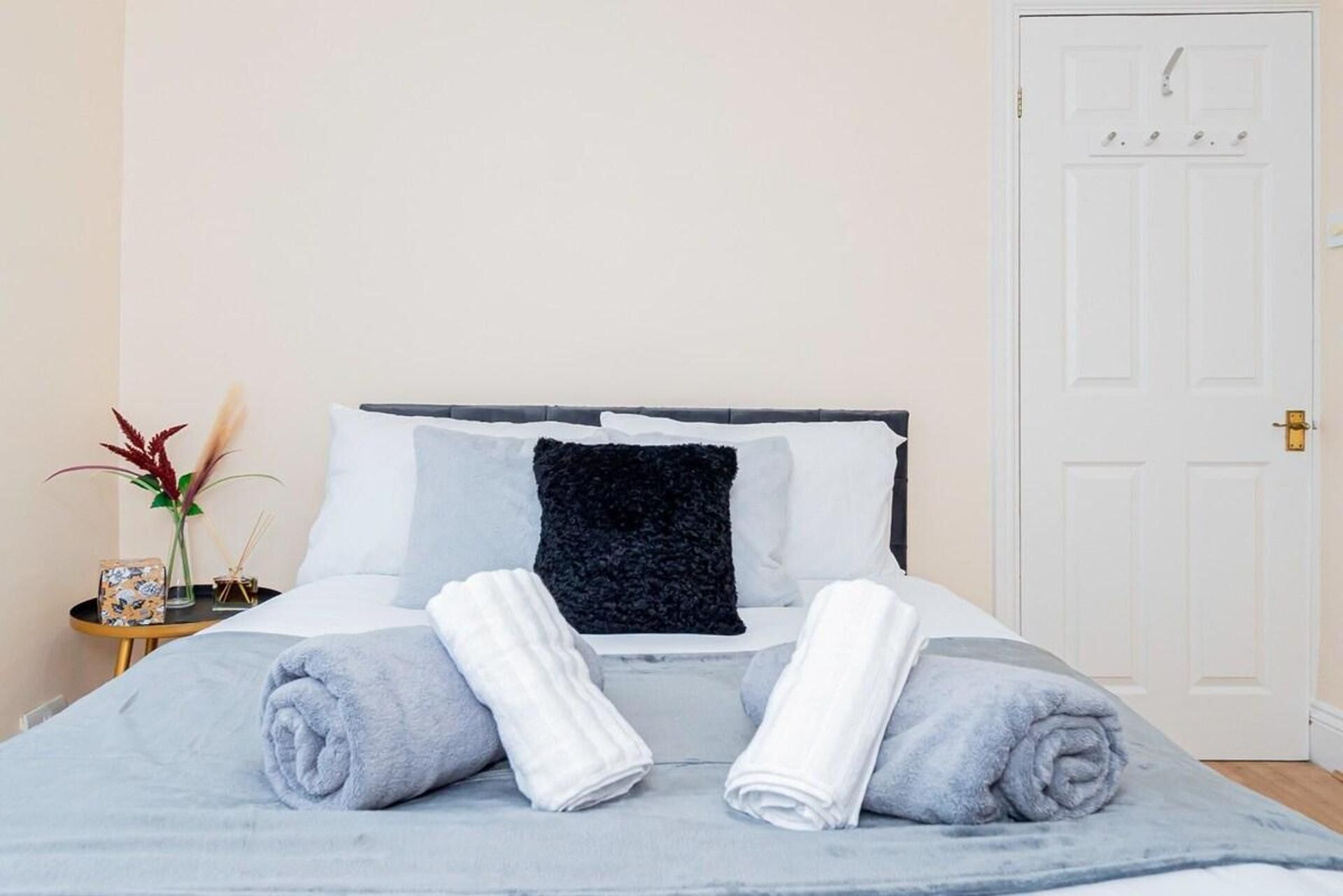 A neatly made bed with a grey blanket, two rolled towels, and a dark pillow.