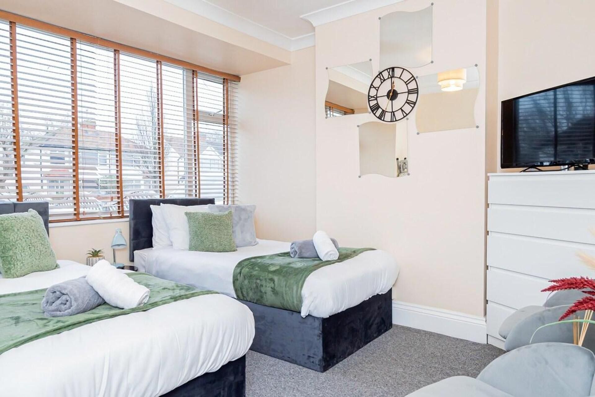 A hotel room with two beds, a large wall clock, a flat-screen TV, and a view of the city through the windows.