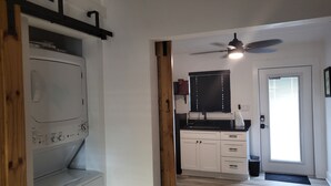 Une buanderie équipée d’un lave-linge et d’un sèche-linge, d’un ventilateur de plafond, d’un coin cuisine avec un plan de travail noir et des placards blancs, et une porte sur laquelle est apposé un panneau « Il est interdit de fumer ».