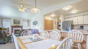 A spacious living room with a fireplace, a dining area with a wooden table and white chairs, and a kitchen with white cabinets and modern appliances.