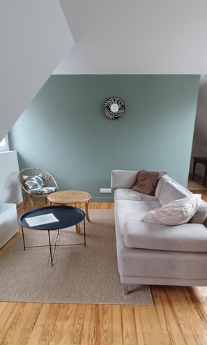 Een woonkamer met een lichtkleurige bank, een rieten stoel, een klein rond tafeltje en een decoratief object aan de muur.