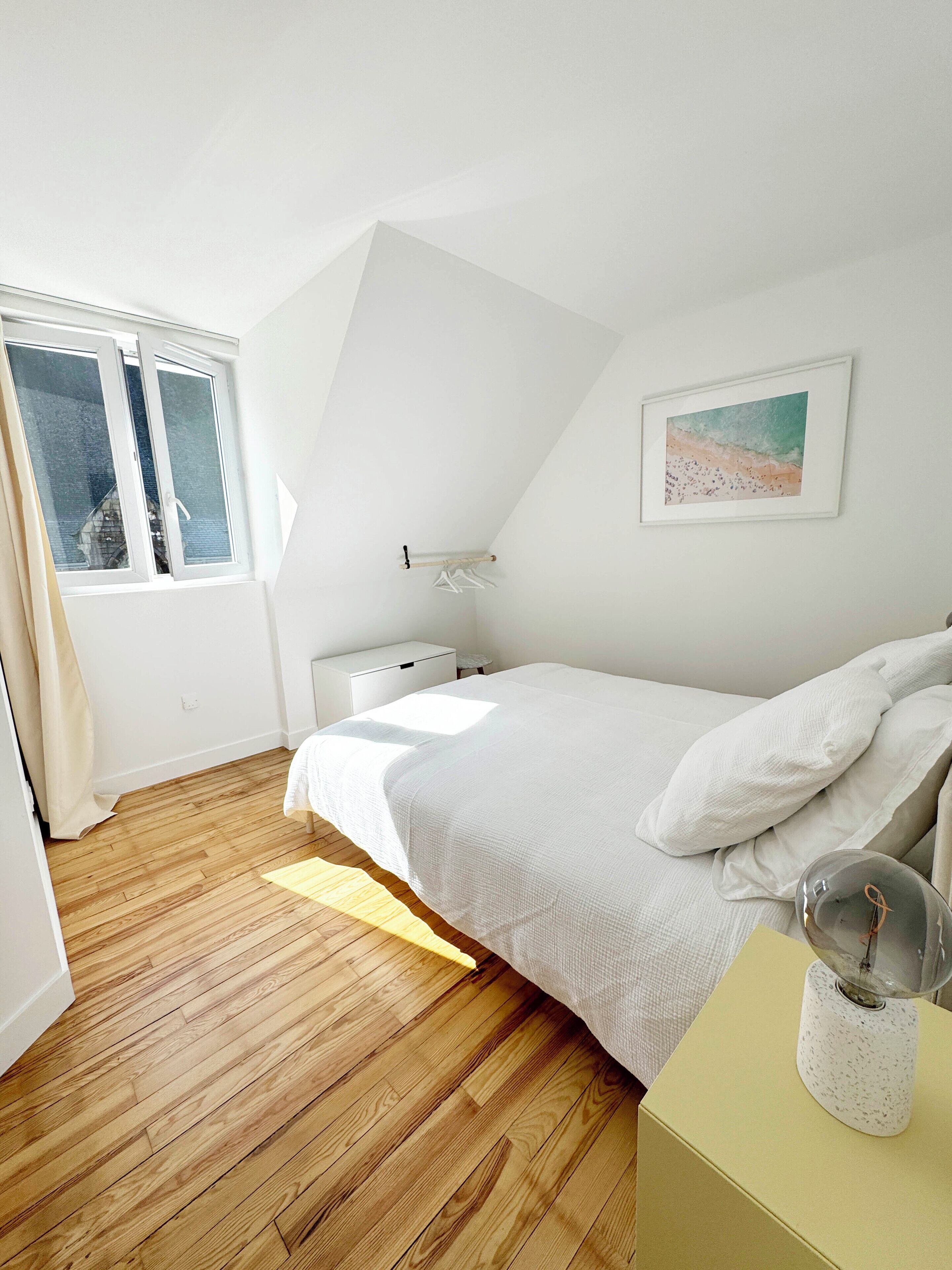 Een slaapkamer met een houten vloer, een bed met wit beddengoed, een nachtkastje, een lamp en een raam met gordijnen.