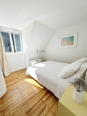 Een slaapkamer met een houten vloer, een bed met wit beddengoed, een nachtkastje, een lamp en een raam met gordijnen.