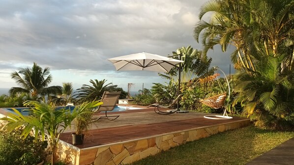 A poolside area with a wooden deck, lounge chairs, and a hanging chair. - Vacances L'ouest Reunion (La Possession)