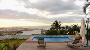 A pool with two lounge chairs and a white umbrella, overlooking a coastal cityscape with a view of the sea and industrial structures. - Vacances L'ouest Reunion (La Possession)