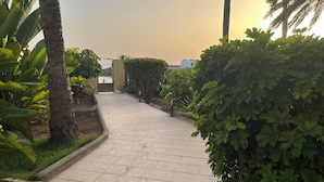 Un chemin pavé, bordé d’une végétation luxuriante et de palmiers, mène à une entrée fermée par un portail.