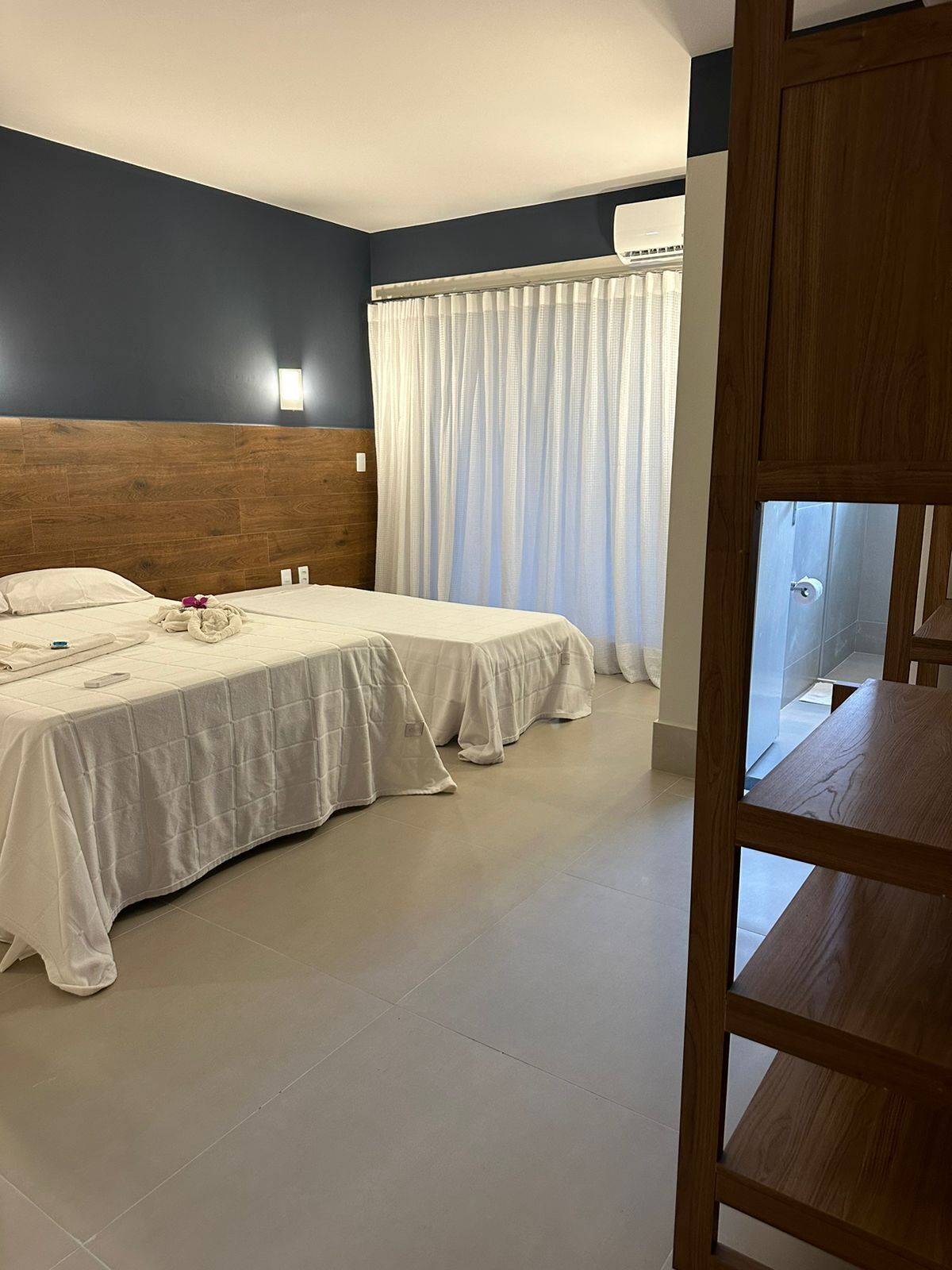 A hotel room with two beds, a wooden headboard, a window with white curtains, and a wooden door.