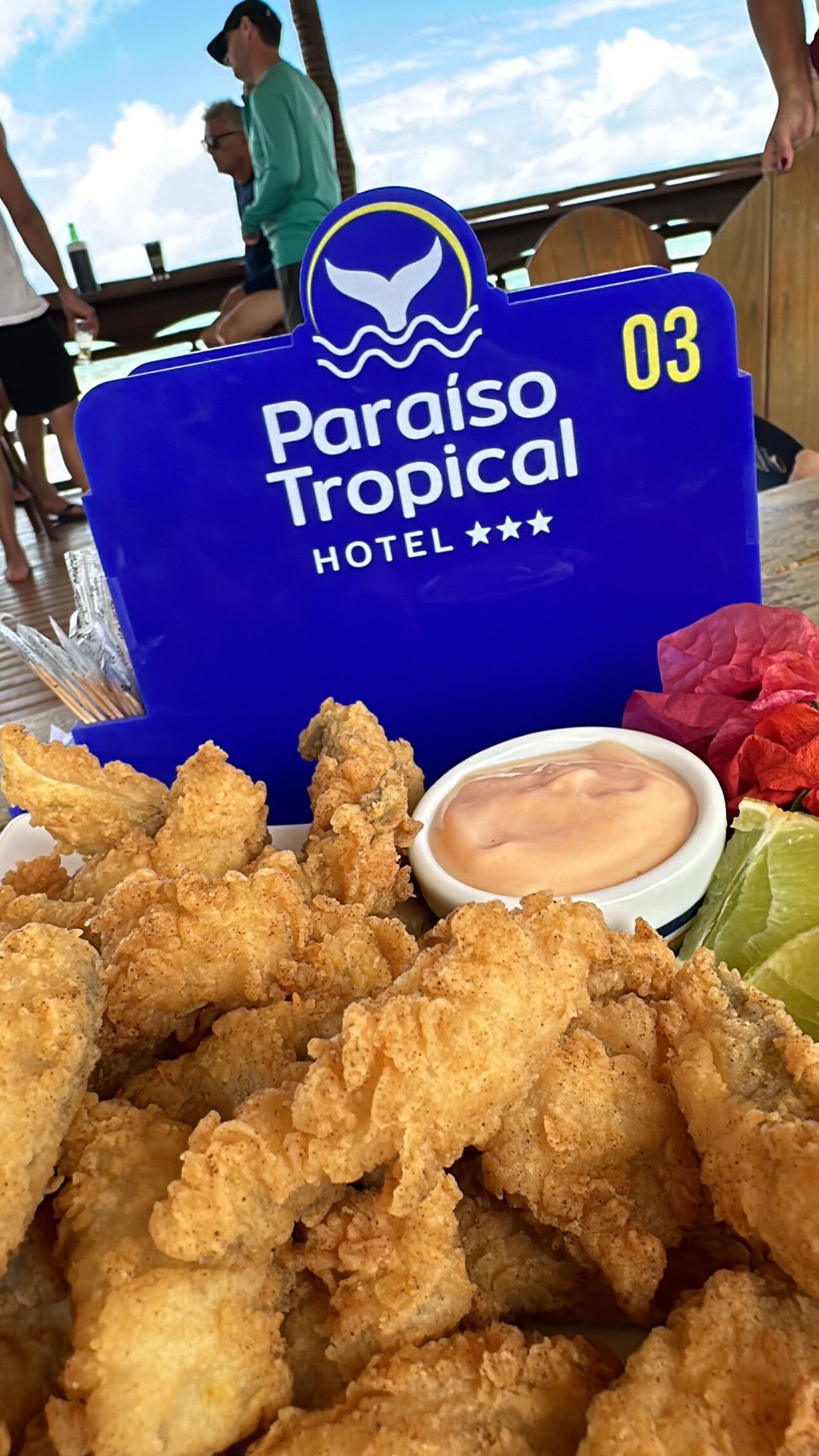 Un letrero azul con el logotipo y el nombre del Hotel Paraíso Tropical, número 03, y un plato de pollo frito con salsa y limón.