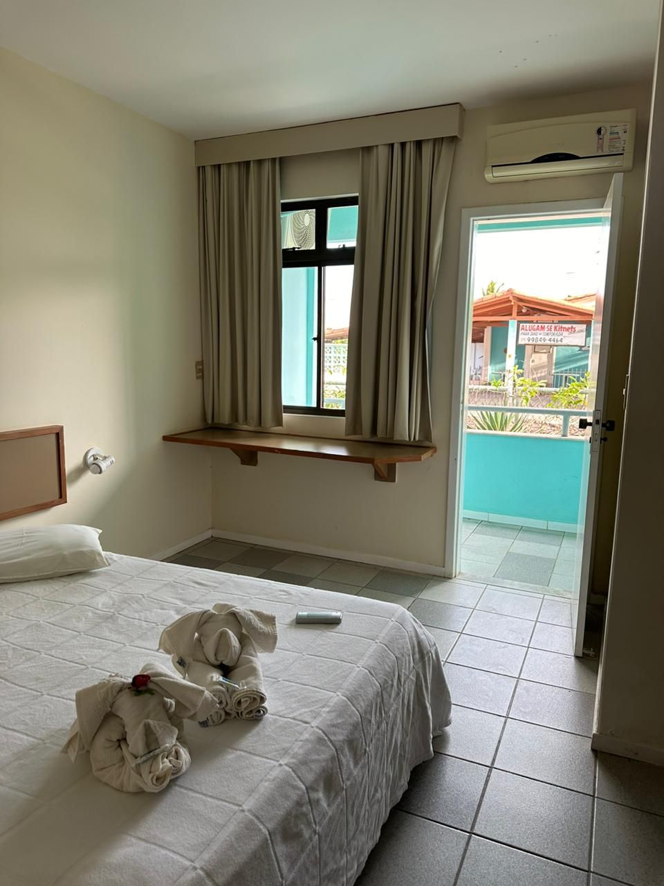 A hotel room with a bed, a window with curtains, a small balcony, and an air conditioning unit.