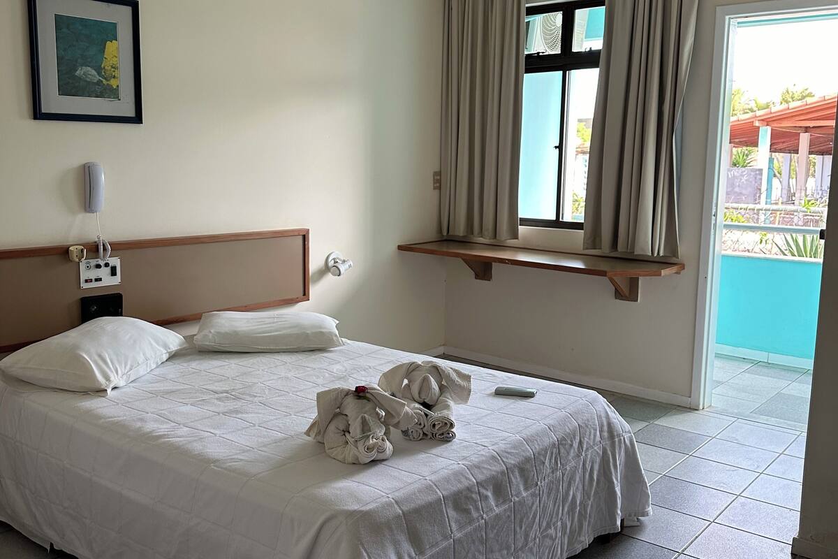 Habitación de hotel con cama, teléfono en la mesita de noche, ventana con cortinas y vistas al exterior.