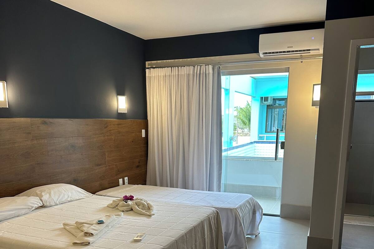 Habitación de hotel con dos camas, cabecero de madera, un ventanal grande con cortinas y un equipo de aire acondicionado.