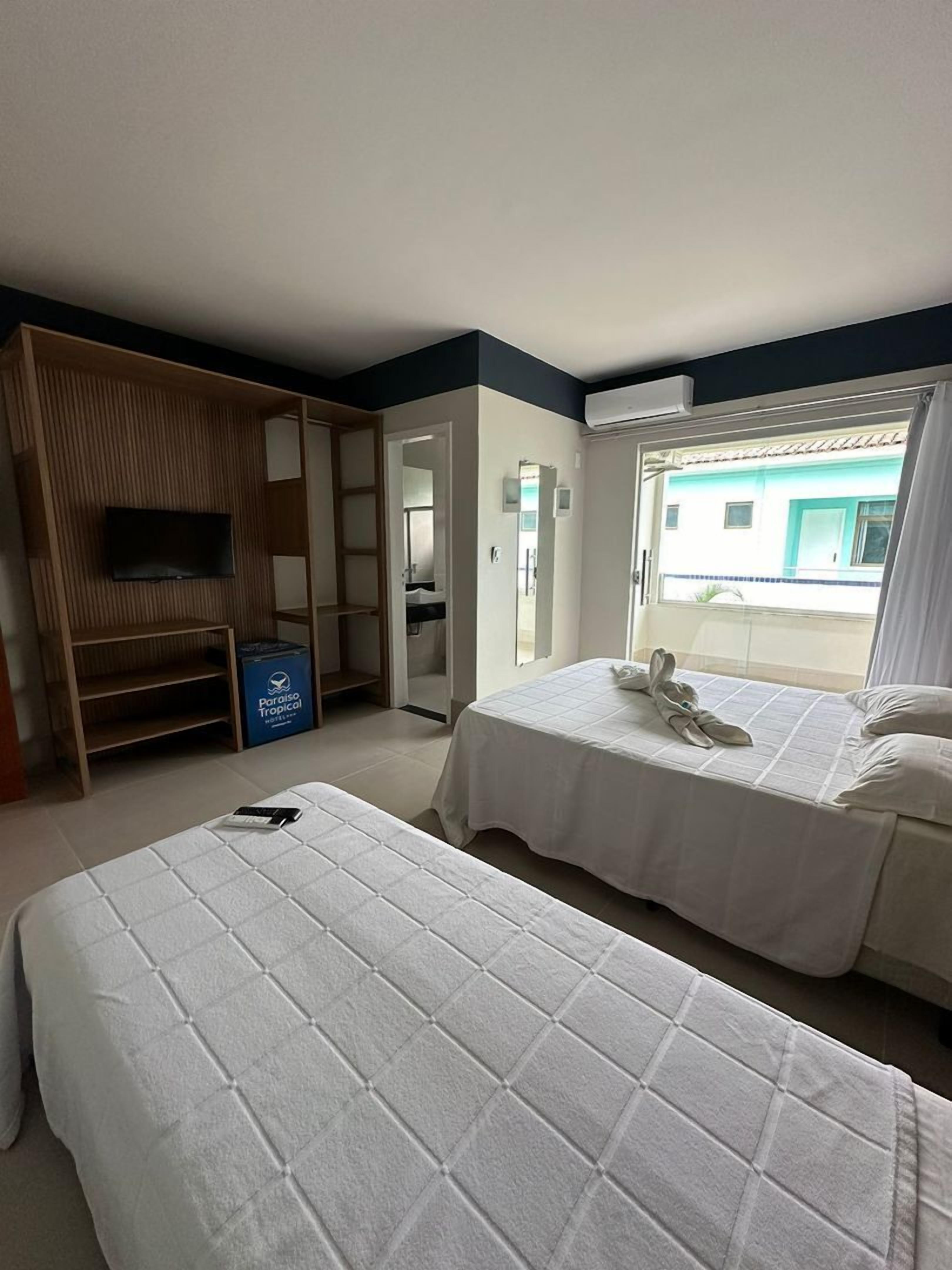 A hotel room with two beds, a TV, a wardrobe, and a view of a building outside.