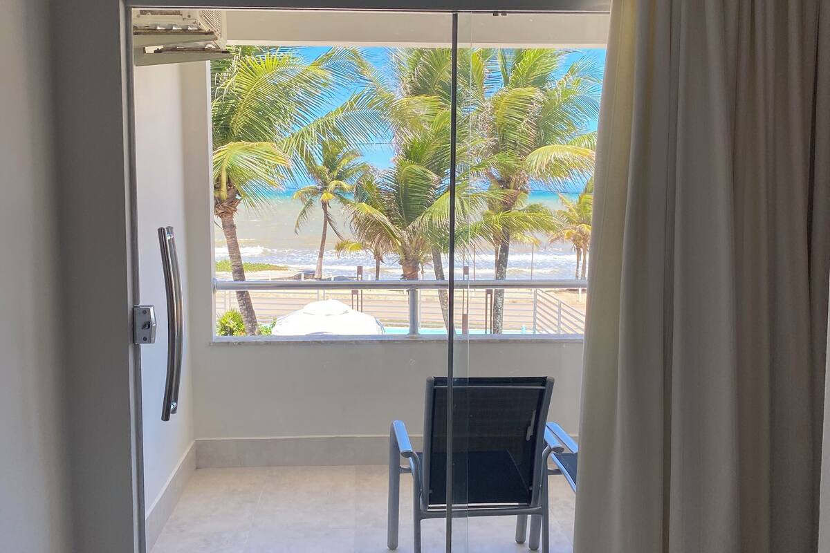 Una habitación con puerta corrediza de vidrio que da a un balcón con vista a palmeras y una playa.