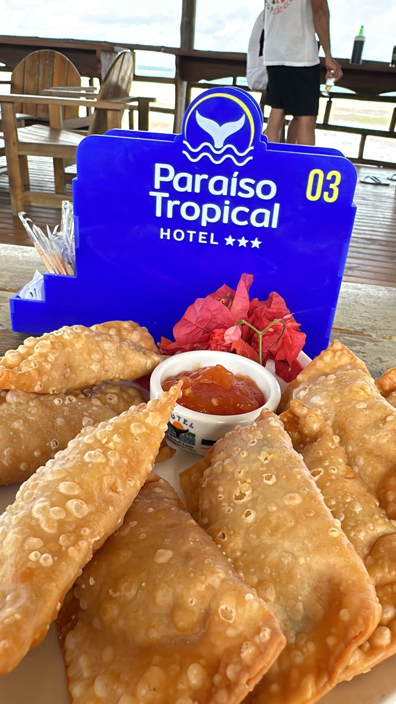 Een bord met gefrituurde gebakjes en dipsaus, een blauw bord met daarop Paraíso Tropical Hotel, en een houten terras op de achtergrond.