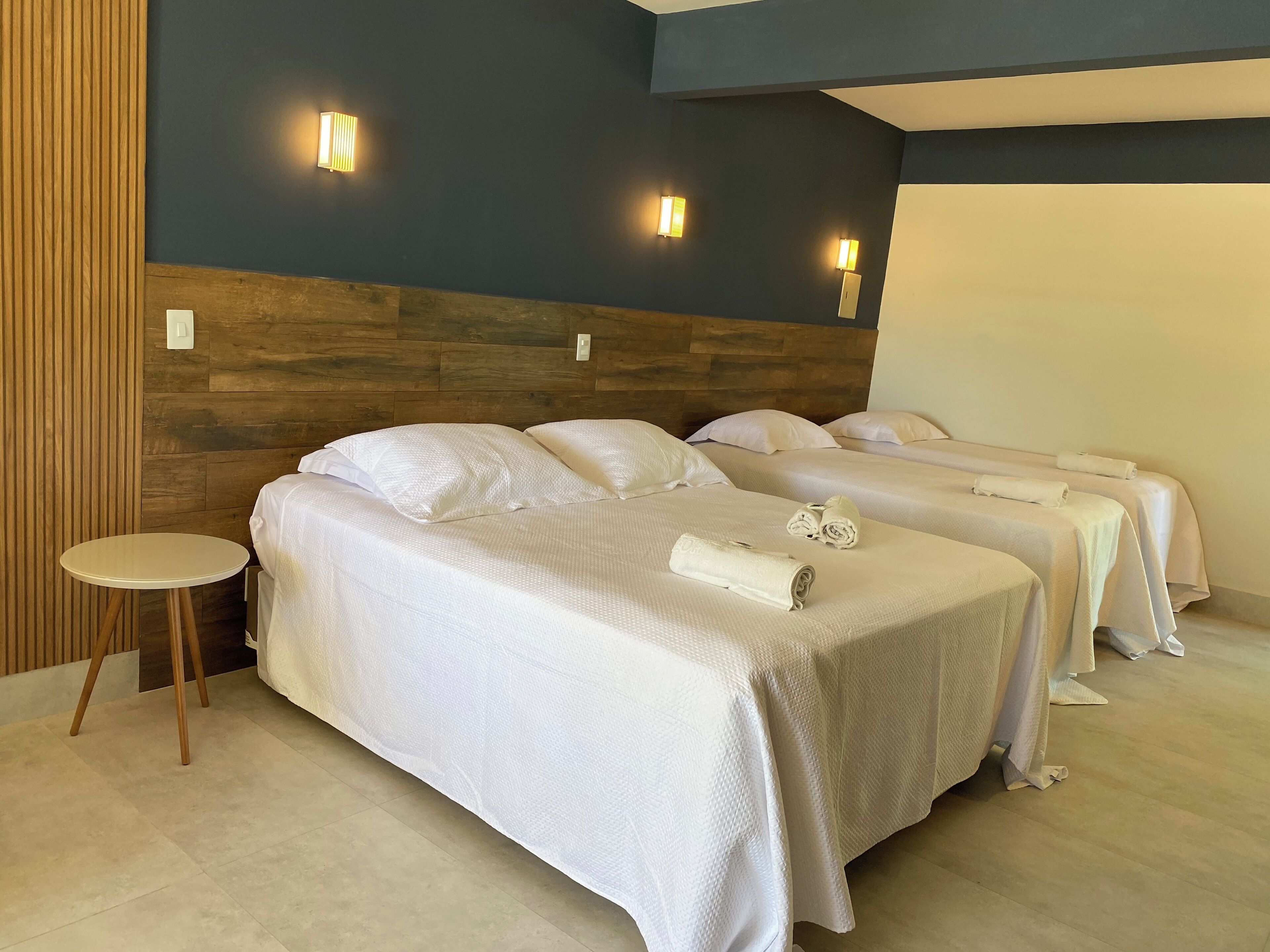 Habitación de hotel con tres camas, cabecero de madera y luces fijadas a la pared.
