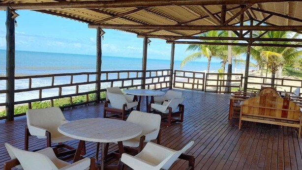 A covered outdoor seating area with a wooden deck, white chairs, a round table, and a wooden bench. - Hotel Paraiso Tropical (Alcobaca)