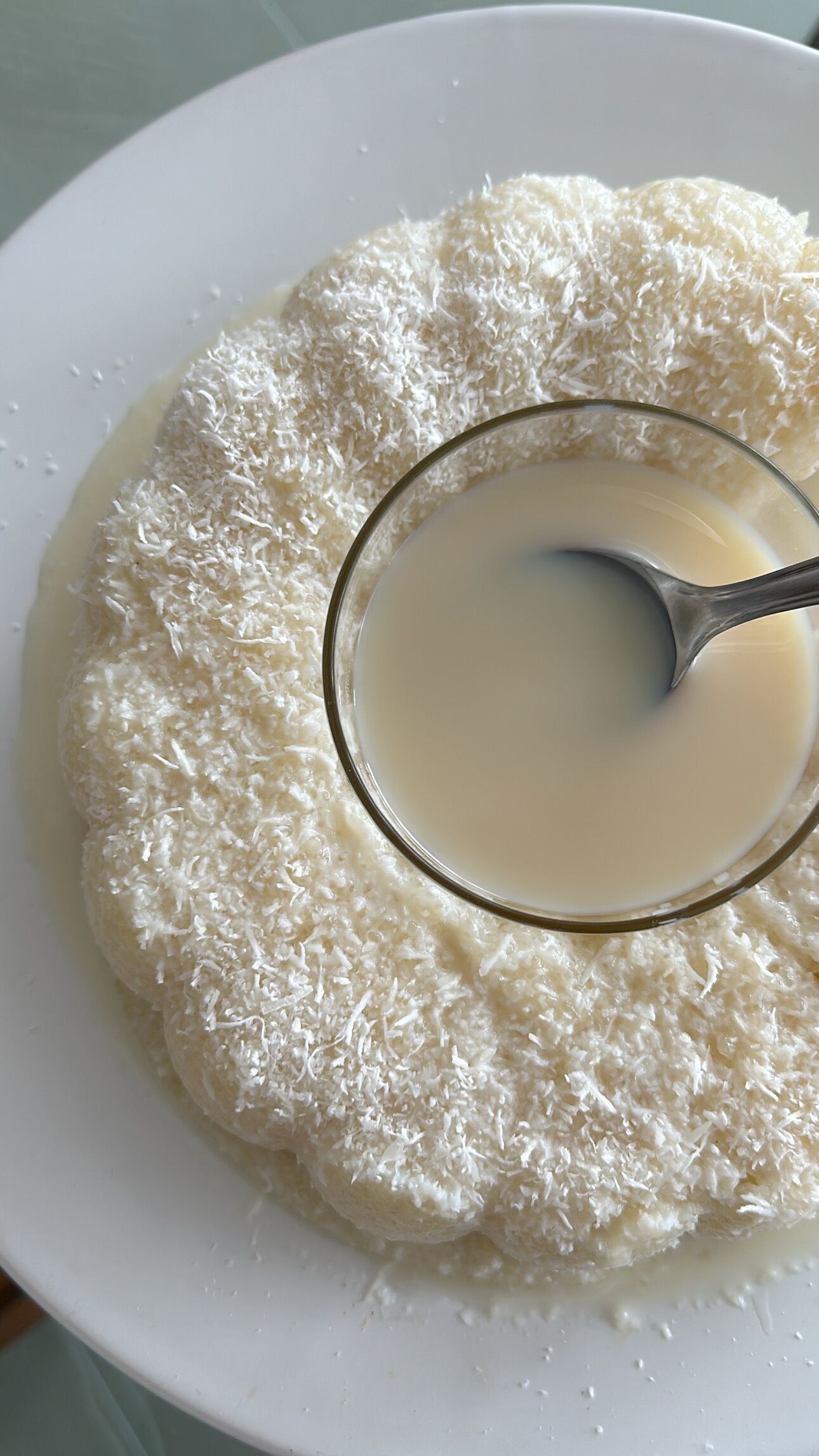 A white plate with a dessert covered in shredded coconut and a glass of milk.