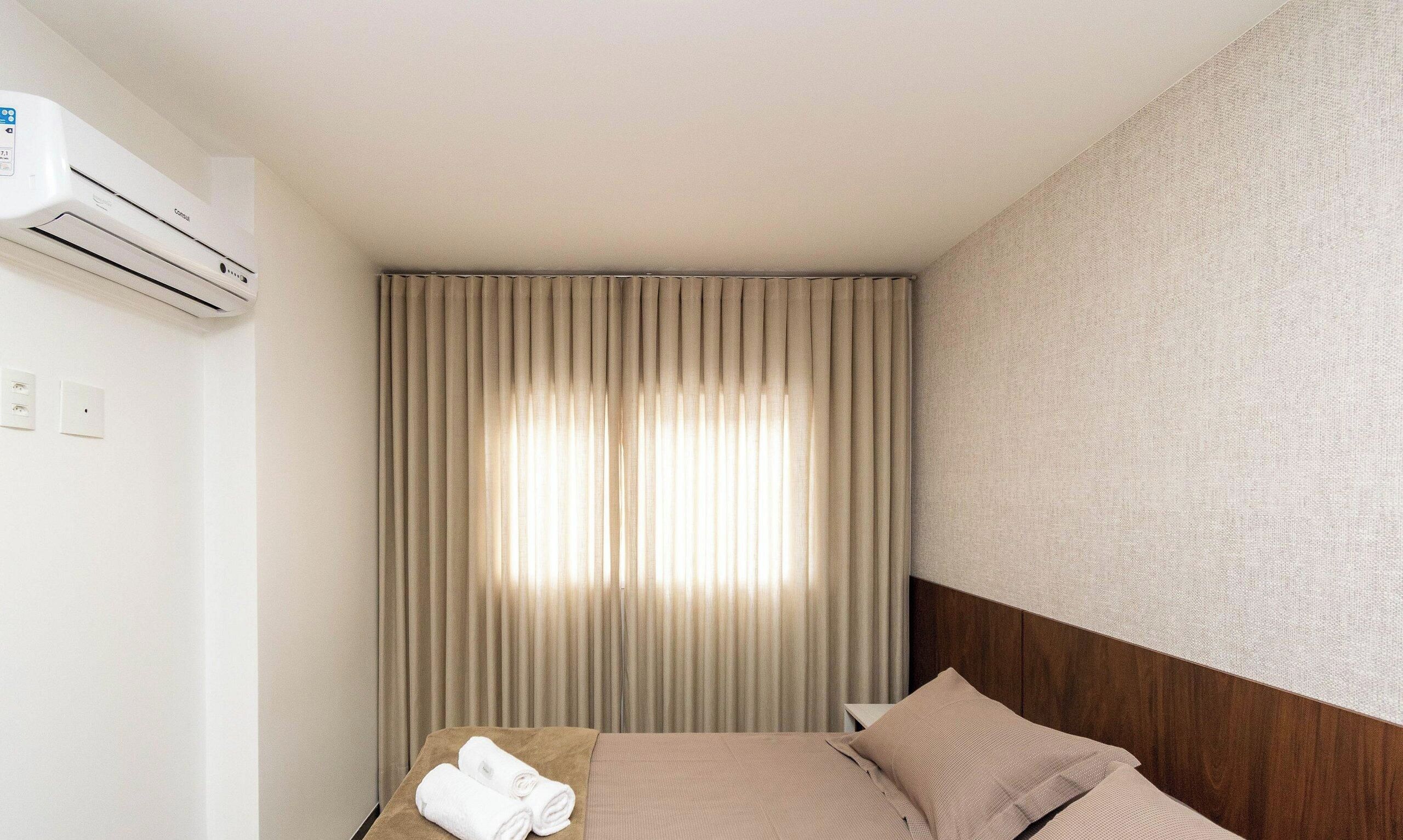 Una habitación de hotel con cama, ventana con cortinas, equipo de aire acondicionado e interruptor de luz en la pared.