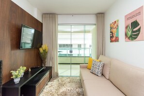 A living room with a flat-screen TV mounted on a wooden wall, a beige sofa with patterned cushions, a glass door leading to a balcony, and two framed artworks on the wall. - Ap 2q St Bueno Ed Pontal Premium PPB1206 (Goiânia)