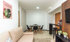 Ein Wohnzimmer mit einer Couch, einem Esstisch mit Stühlen, einer Küchenzeile mit einer Theke und Hocker sowie einem an der Wand befestigten Fernseher.