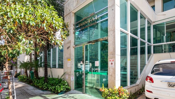 Un edificio moderno con entrada de vidrio, un coche blanco estacionado afuera y un árbol a la izquierda.