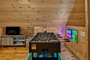 A room with a foosball table, a television, and a neon sign indicating a game room. - Rustic Ridge 3-Level Log Cabin, Covered Deck, Hot Tub, Adult Retreat, Kids Loft (Ellijay)