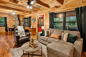 A cozy cabin interior with a wooden ceiling, a fireplace, a leather armchair, a sofa, and large windows offering a view of the outdoors.