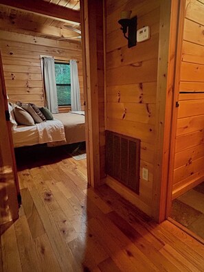 A wooden cabin interior with a bed, a window with curtains, a wall-mounted heater, and a wooden floor. - Rustic Ridge 3-Level Log Cabin, Covered Deck, Hot Tub, Adult Retreat, Kids Loft (Ellijay)