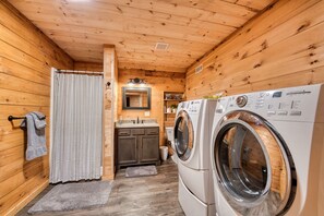 A compact, wooden-floored laundry room with two front-loading washing machines, a small sink, and a mirror above it. - Rustic Ridge 3-Level Log Cabin, Covered Deck, Hot Tub, Adult Retreat, Kids Loft (Ellijay)