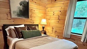 Habitación con paneles de madera, una cama, una mesita de noche, una lámpara y una ventana con cortinas.