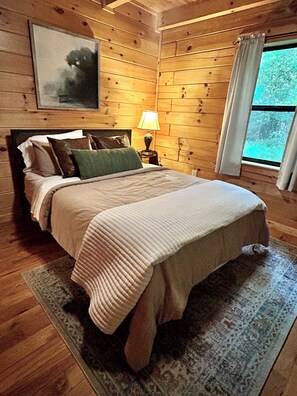 A wooden-paneled room with a bed, a nightstand, a lamp, and a window with curtains. - Rustic Ridge 3-Level Log Cabin, Covered Deck, Hot Tub, Adult Retreat, Kids Loft (Ellijay)