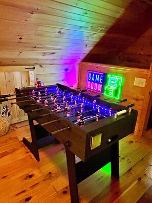 A foosball table in a room with wooden walls and a neon sign that reads 'GAME ROOM'.