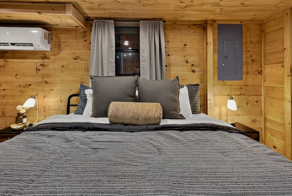 A wooden room with a bed, two bedside lamps, a window with curtains, and a wall-mounted air conditioner. - Rustic Ridge 3-Level Log Cabin, Covered Deck, Hot Tub, Adult Retreat, Kids Loft (Ellijay)