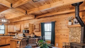 A cozy cabin-style kitchen with wooden walls, a stone fireplace, and a dining area with a round table and chairs. - Rustic Ridge- A cozy 2-bedroom+loft cabin outside Elijay w/hottub, firepit. (Ellijay)