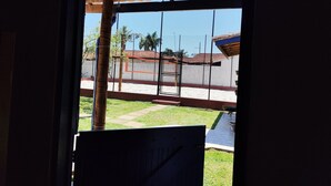 A view through a window with a metal gate, a grassy yard, and a building with a red roof in the background. - House with sand court 100m from the beach (Praia da Boracéia São Sebastião)