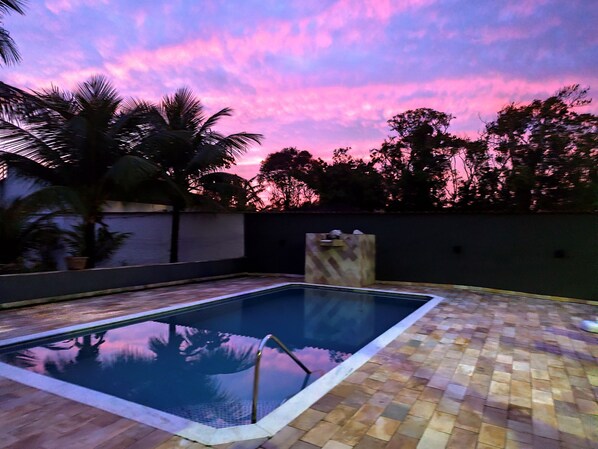 Una piscina rectangular con un borde de baldosas, rodeada por un muro bajo y palmeras.