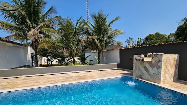 A swimming pool with clear blue water, surrounded by palm trees and a stone wall. - House with sand court 100m from the beach (Praia da Boracéia São Sebastião)