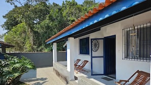 A covered outdoor area with a tiled floor, two wooden lounge chairs, a concrete bench, and a wall with a blue door and window. - House with sand court 100m from the beach (Praia da Boracéia São Sebastião)
