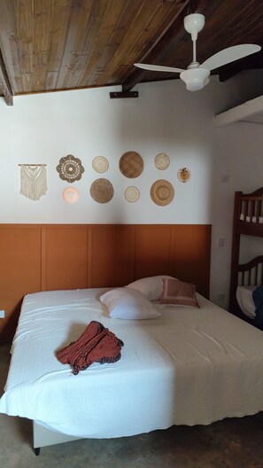 A bedroom with a wooden headboard, a bed with white bedding, a red blanket, and a ceiling fan. Decorative wall hangings are visible above the bed.