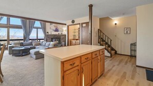A spacious living area with a large window, a fireplace, a kitchen with wooden cabinets, and a staircase leading to another level. - Pocono Mountains Camelback Retreat-Stunning Views (Tannersville)