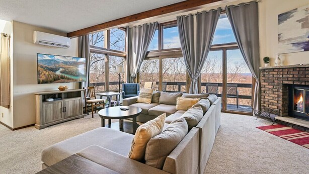 A cozy living room with a sectional sofa, a fireplace, and large windows offering a view of the outdoors. - Pocono Mountains Camelback Retreat-Stunning Views (Tannersville)