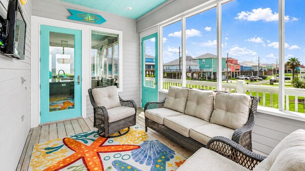 A covered porch with wicker furniture, a starfish rug, and a view of houses. - Beach View 4BR | Sleeps 14 | Fenced Yard (Crystal Beach)