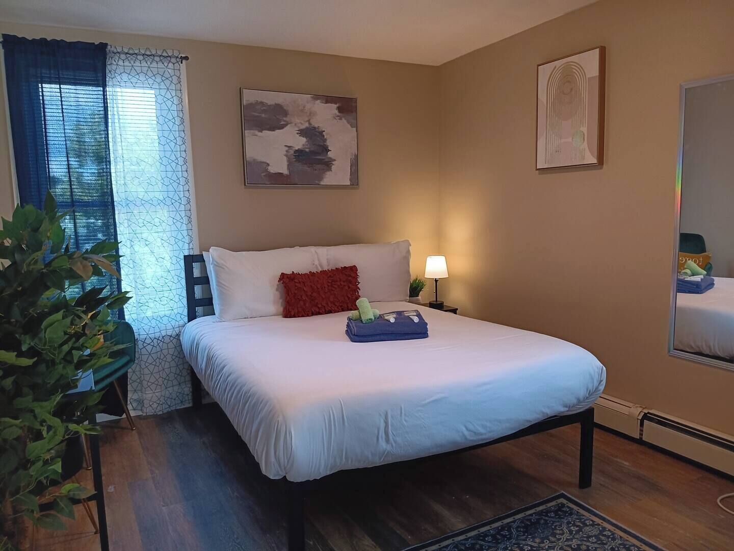 A hotel room with a bed, a nightstand, a lamp, a plant, and artwork on the walls.