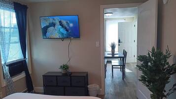 Una camera da letto con un letto, una cassettiera, una TV a parete, una pianta in vaso e vista su una sala da pranzo.