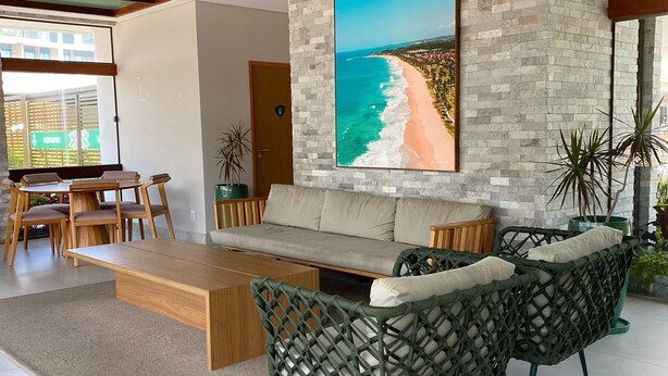 A modern living room with a stone accent wall, a large beach mural, a sofa, two green woven chairs, and a wooden coffee table. - Mana Beach Experience 217Aloha byLaPlaya (Ipojuca)