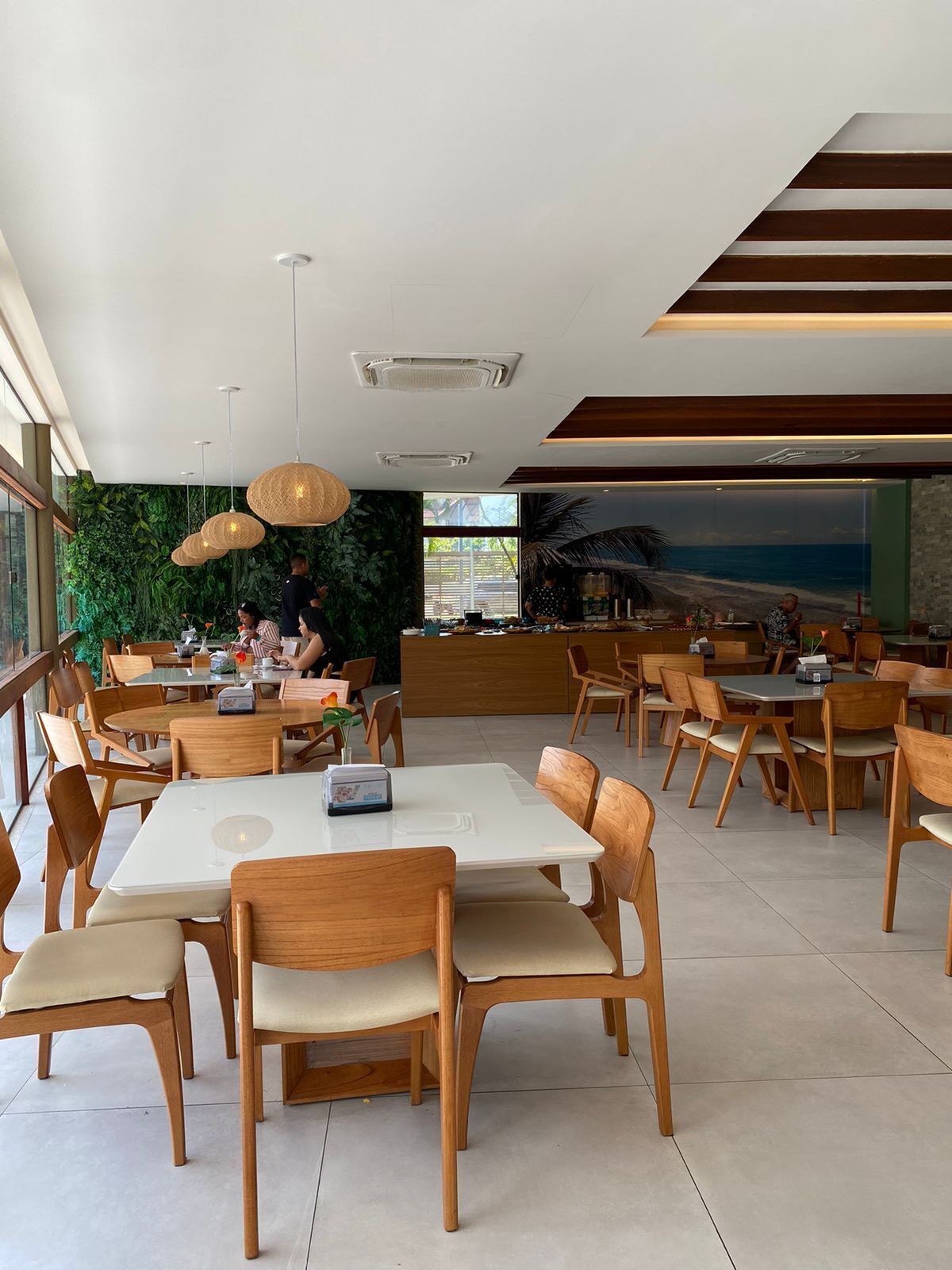 A modern restaurant with wooden tables and chairs, a green wall with plants, and a view of the ocean through large windows.