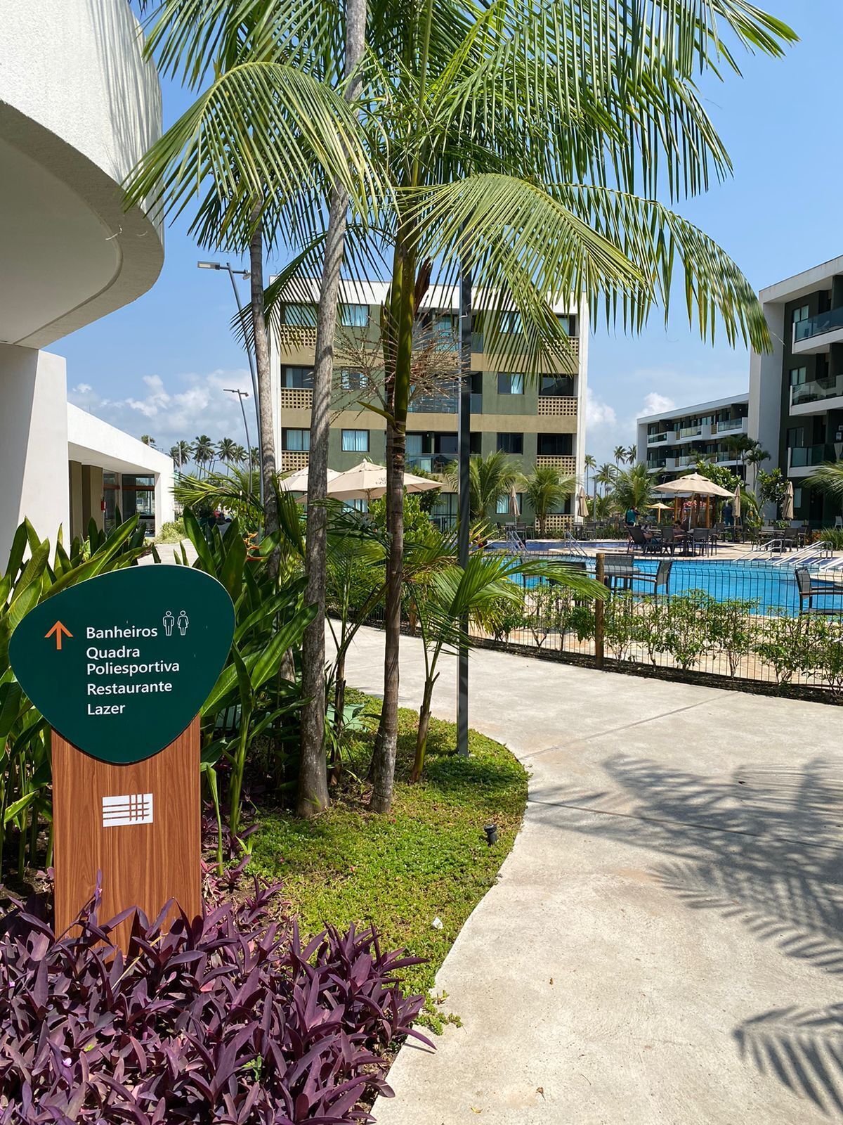 Un hotel con piscina, palmeras y un cartel que indica servicios como restaurante y centro deportivo.
