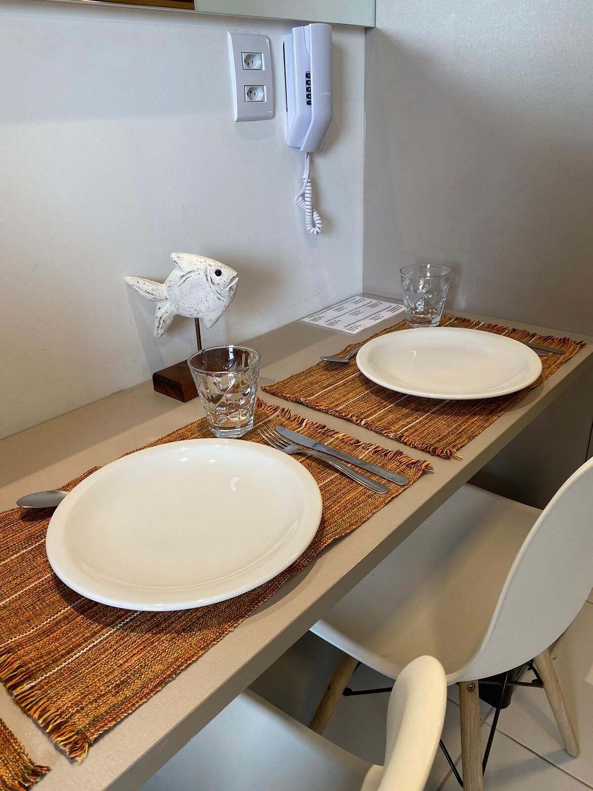 A dining table with two white plates, a fork, a knife, and a glass. There is a white fish-shaped decoration on the wall and a telephone on the wall.