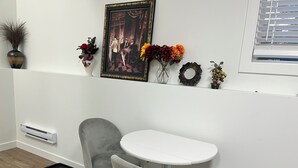 Una habitación con pared blanca, un cuadro enmarcado, una mesita, dos sillas, un radiador y una ventana con persianas.