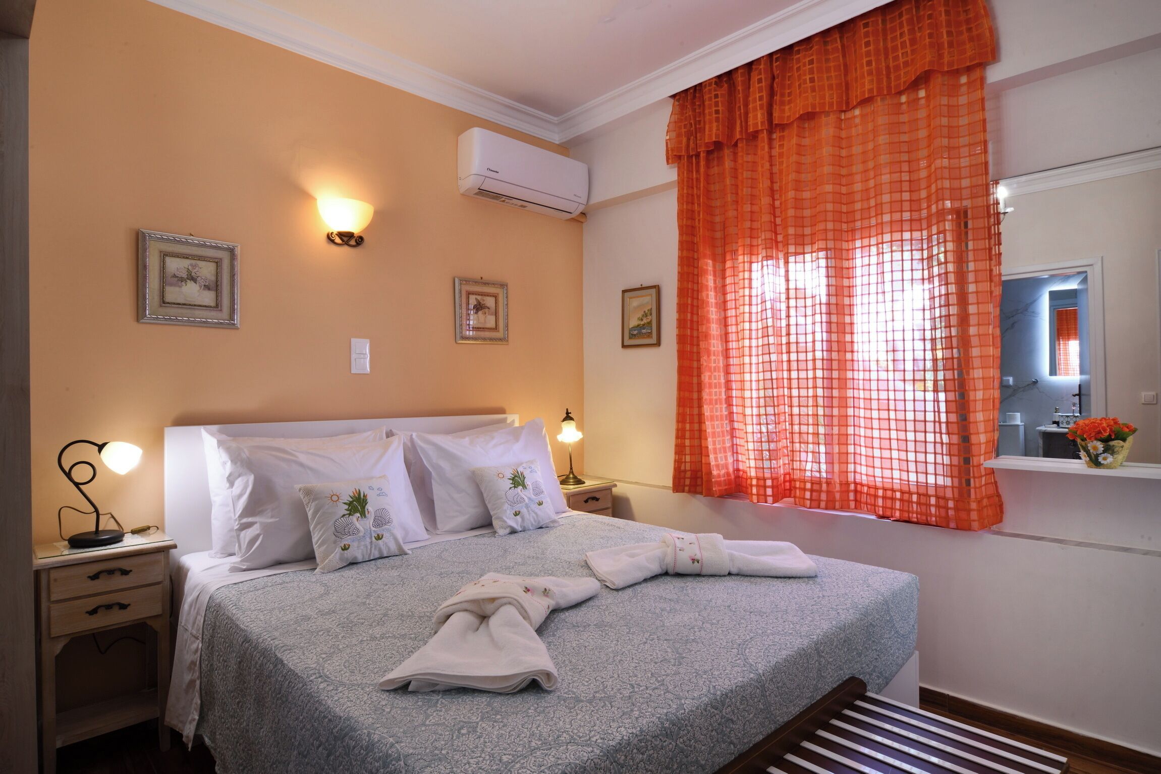 A neatly made bed with white linens and decorative pillows, a bedside lamp, and a window with orange curtains.